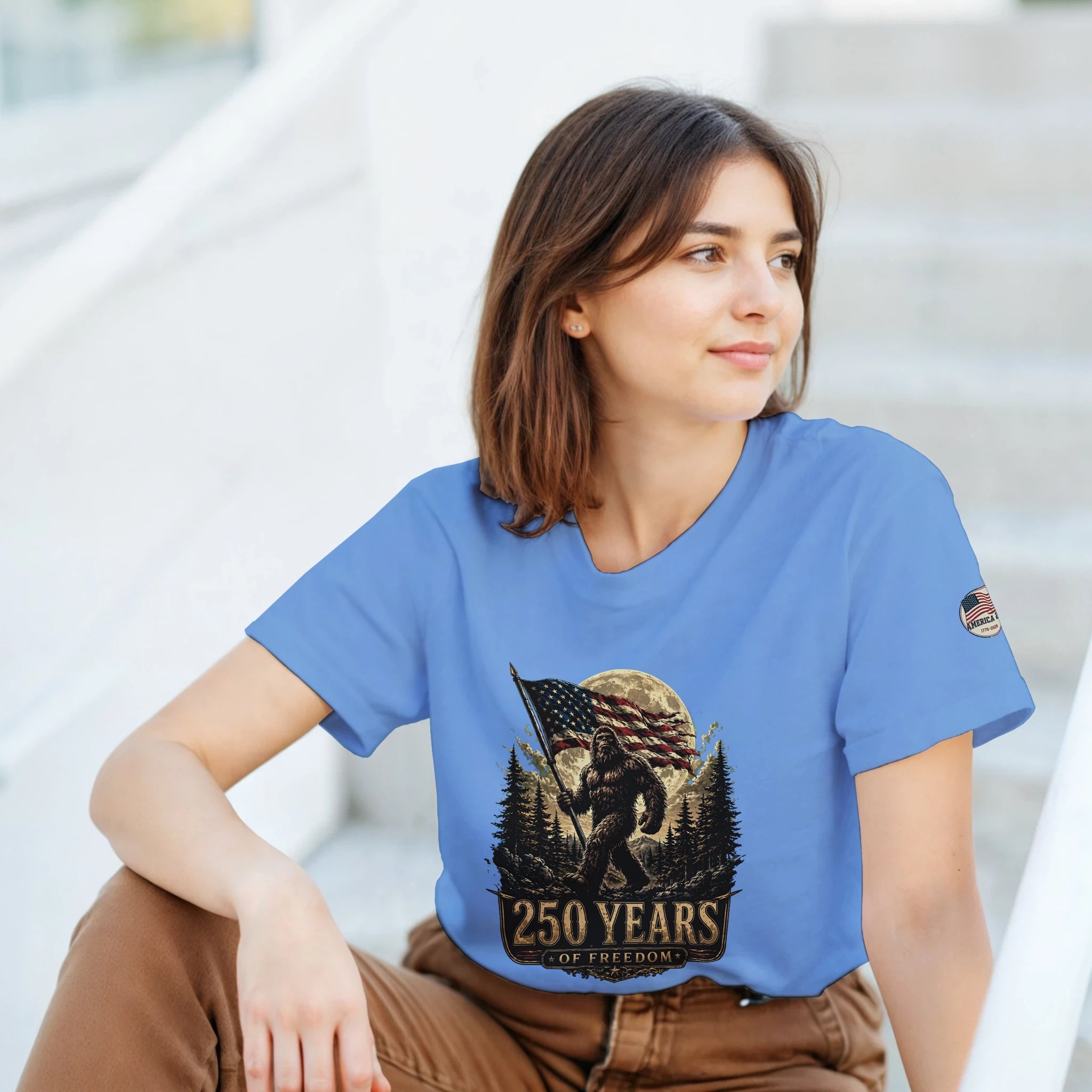 Woman wearing a blue t-shirt with a Bigfoot graphic design and text, sitting outdoors.